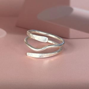 Peut inclure: Une bague enroulante argentée avec une texture martelée. La bague présente un design en spirale avec trois bandes distinctes qui s'enroulent autour du doigt. La bague est présentée sur une surface rose, avec des ombres douces en arrière-plan.