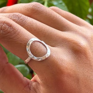 Peut inclure: Une bague en argent avec un design ovale ouvert. La bague est portée au doigt, avec une surface texturée sur l'ovale. Le fond présente un feuillage vert et des fleurs rouges.