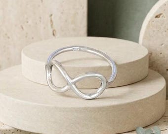 May include: A silver-colored infinity symbol ring. The ring is displayed on a beige, circular platform. The ring's design features a continuous loop, symbolizing eternity. The background includes additional platforms and a green backdrop.