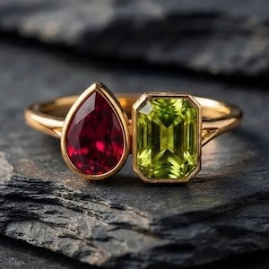 May include: A gold ring featuring two prominent gemstones: a deep red pear-cut stone and a bright lime green emerald-cut stone. The stones are set side-by-side on a thin gold band, creating a distinct contrast between the two faceted shapes.