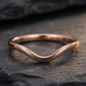 May include: A polished rose gold ring featuring a smooth, curved V-shaped band. The metal has a reflective, metallic finish with a consistent, rounded profile throughout the entire circumference of the jewelry piece, showcasing a simple design.