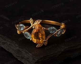 May include: A gold ring featuring a pear-shaped, amber-colored gemstone as the centerpiece, flanked by two smaller, light blue gemstones. The ring's band has a delicate, leaf-like design. The ring is set against a dark background.