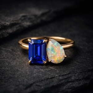 May include: A gold ring featuring a large, rectangular, deep blue gemstone and a pear-shaped opal with iridescent flashes of color. The ring is set against a dark, textured background, highlighting the gemstones' brilliance.