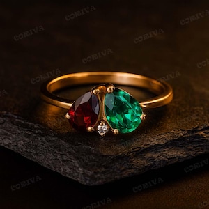 Könnte beinhalten: Ein goldener Ring mit zwei tropfenförmigen Edelsteinen, einem tiefroten und einem smaragdgrünen. Zwischen den beiden größeren Steinen befindet sich ein kleiner, runder Diamant. Der Ring liegt auf einer dunklen, strukturierten Oberfläche.