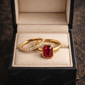 May include: Two gold rings in a black ring box. One ring features a rectangular, deep red gemstone. The other ring is a gold band with small, clear gemstones. The box is open, revealing a beige interior.