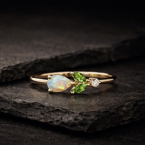May include: A gold ring with a pear-shaped opal, two green peridot leaves, and a small diamond. The ring is set against a dark, textured background. The opal and peridot are the focal point of the ring's design.