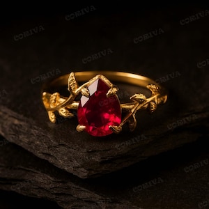 Puede incluir: Un anillo de oro con una piedra preciosa roja en forma de pera como pieza central. La banda presenta un diseño orgánico y texturizado que recuerda a ramas y hojas delicadas que se extienden para sujetar la piedra central en su engaste.