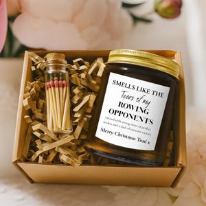 May include: A candle in a glass jar with a gold lid, inside a cardboard box with shredded paper. The candle has a label that reads "SMELLS LIKE THE Tears of my ROWING OPPONENTS". A small glass vial of matches is also in the box.