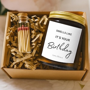 May include: A birthday gift set in a brown box, featuring a candle with a gold lid and a label saying "Smells Like It's Your Birthday." A small glass vial holds red-tipped matches.