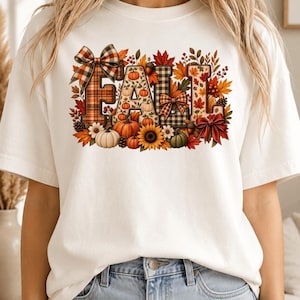 Könnte beinhalten: Ein weißes Kurzarm-T-Shirt mit einem Grafikdruck des Wortes FALL in großen Blockbuchstaben. Die Buchstaben sind mit Karomustern, Kürbissen, Sonnenblumen, Herbstblättern, Tannenzapfen und dekorativen Schleifen in Orange und Braun verziert.