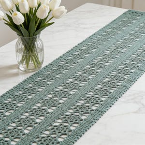 Può includere: Un runner da tavola rettangolare all'uncinetto color verde salvia caratterizzato da un intricato motivo a pizzo ripetuto con dettagli geometrici traforati e fasce verticali piene, come lungo elemento tessile decorativo.