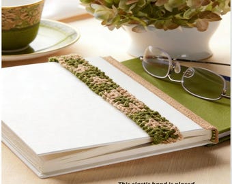 May include: A white book with a green and tan crocheted elastic band bookmark. The bookmark is placed around the book to mark the page. A pair of glasses and a cup and saucer are in the background.