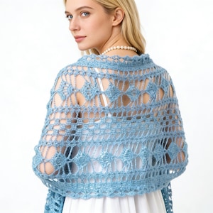 Op de afbeelding: Een lichtblauwe, open geweven gehaakte sjaal met een ruitpatroon. De sjaal wordt over een witte jurk gedragen. De sjaal heeft een delicaat, kantachtig ontwerp, ideaal om in laagjes te dragen. Het model kijkt weg van de camera.