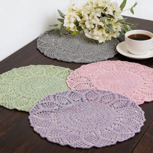 May include: Four crocheted placemats in shades of gray, green, pink, and lavender are displayed on a dark wood surface. A white teacup and saucer with coffee and a bouquet of white flowers are also on the table. The placemats have a circular, openwork design.