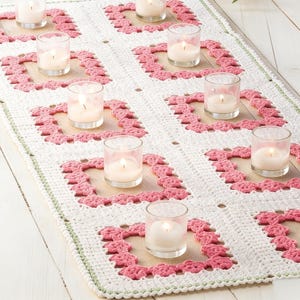May include: A white crocheted table runner with a pink square design. Clear glass votive candle holders with lit candles are placed on the runner. The runner is on a light-colored wooden surface.