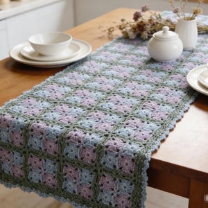 May include: A handmade crochet table runner in a granny square pattern. The runner features squares of blue, lavender, and green yarn. The table runner is displayed on a wooden table with white dishes and a sugar bowl.