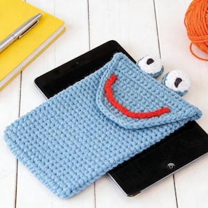May include: A light blue crochet tablet case with a red stitched smile, on a black tablet. Two small, white and blue crochet eyes are beside the case. A yellow notebook, silver pen, and orange ball of yarn are in the background.
