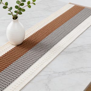 Könnte beinhalten: Rechteckiger Tischläufer mit strukturiertem Wabenmuster. Der Läufer hat Streifen in Braun, Grau und Creme. Eine weiße Vase mit grünen Zweigen steht auf dem Tisch. Der Tisch hat eine marmorähnliche Oberfläche.