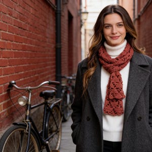 Puede incluir: Una persona con un abrigo gris, un cuello alto blanco y una bufanda calada de color óxido. La bufanda está drapeada alrededor del cuello. Una bicicleta negra está aparcada contra una pared de ladrillos rojos.
