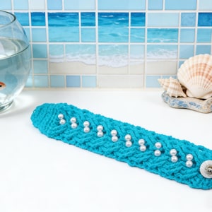 May include: A turquoise blue, knitted headband with white pearl accents and a white button closure. The headband is displayed on a white surface, with a fishbowl and seashells in the background.