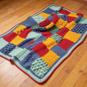 May include: A vibrant, hand-crocheted blanket with a patchwork design. The blanket showcases squares in red, yellow, blue, and green, with diverse textured stitches. A red border and a light blue trim frame the blanket, creating a cosy and colourful appearance.