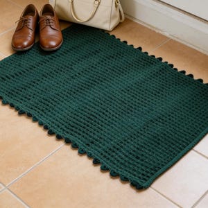 May include: A dark green rectangular doormat with a textured, grid-like pattern and a scalloped edge. Brown leather shoes and a cream-colored handbag are placed near the mat, suggesting an entryway setting.