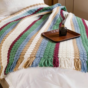 Puede incluir: Una manta de ganchillo a rayas con flecos. La manta presenta rayas blancas, verdes, azules, burdeos y beige. Una bandeja marrón con un difusor se encuentra sobre la manta, que está sobre una cama.