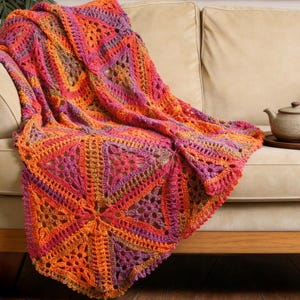 May include: A vibrant, handmade crochet blanket in shades of orange, pink, and purple, draped over a beige sofa. The blanket features a geometric starburst design and intricate openwork patterns, adding a cozy, decorative touch to the living space.