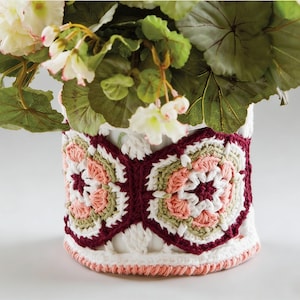 May include: A white cylindrical vase with a crocheted band featuring a geometric pattern in burgundy, peach, and green. The vase is filled with green foliage and white and pink flowers. The crochet work adds a handmade touch.