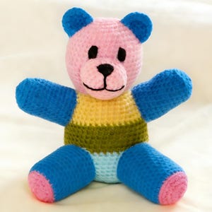 May include: A handmade crochet teddy bear in a sitting position. The bear has a pink head and ears, blue arms and legs, and a striped body in yellow, green, and light blue. The bear's paws are pink, and it has a black stitched mouth and eyes.