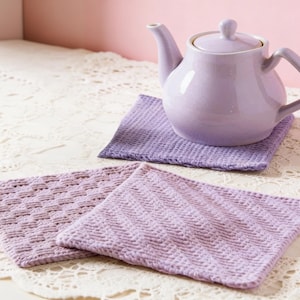 May include: A lavender ceramic teapot with a matching lid sits on a square, textured lavender coaster. Two additional lavender coasters with different woven patterns are in the foreground. The items are on a white lace tablecloth.