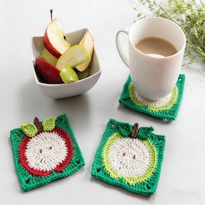 Könnte beinhalten: Eine weiße Tasse mit einem hellbraunen Getränk steht auf einem gehäkelten Untersetzer in Grün, Weiß und Gelb. Zwei weitere Untersetzer mit Apfelmotiven sind zu sehen. Eine Schüssel mit Apfelscheiben ist ebenfalls vorhanden.
