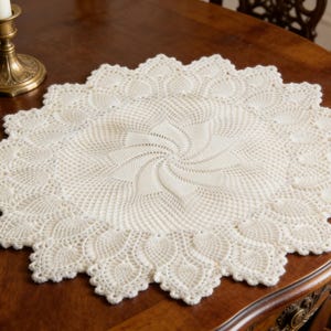 May include: Cream-colored, round, crocheted doily with a scalloped edge. The center features a spiral design. A brass candlestick with a lit white candle sits on a dark wooden table.