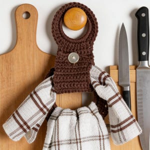 Könnte beinhalten: Ein brauner gehäkelter Küchentuchhalter mit einem Holzknopf und einem silbernen Knopf. Der Halter ist an einem weiß-braunen karierten Küchentuch befestigt. Messer werden auf einem Holzbrett ausgestellt.