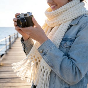 May include: A silver and black film camera is held by a person wearing a light blue denim jacket and a cream-colored, fringed scarf. The background shows a wooden pier extending into the ocean, with the sun setting in the distance.