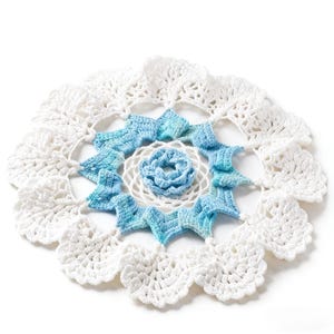 May include: A handmade, circular crochet doily in white and shades of blue. The doily features a central floral design, surrounded by blue star-shaped elements and a scalloped white border. The intricate details highlight the craftsmanship.