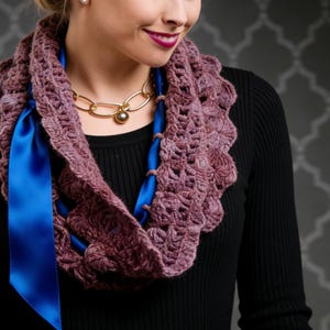 May include: A mauve crochet infinity scarf with a scalloped edge and a bright blue satin ribbon threaded through the design. A gold chain necklace with a pearl pendant is also visible.