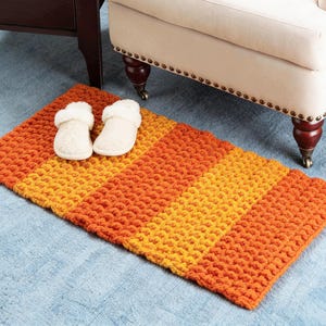 Puede incluir: Una alfombra rectangular tejida a mano en tonos naranja y amarillo, con una superficie texturizada. Dos zapatillas blancas y esponjosas descansan sobre la alfombra. La alfombra está colocada sobre una alfombra azul, cerca de un sillón crema.