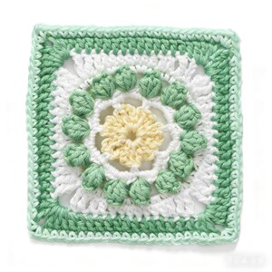 May include: A square, handmade crochet piece featuring a floral design. The center is a pale yellow flower, surrounded by white and green textured stitches. The outer border is a solid green, creating a vibrant, handcrafted look.