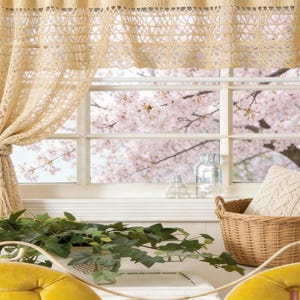 May include: A window with a view of blooming pink cherry blossom trees. Beige crocheted curtains frame the window, tied back with a rope. A woven basket holds a cream-colored pillow. Green plants and yellow furniture are in the foreground.