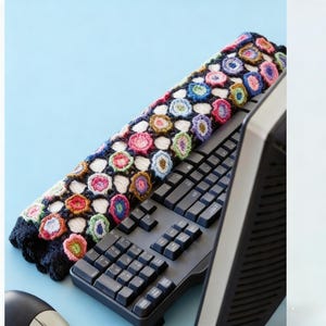 Könnte beinhalten: Eine bunte, gehäkelte Tastaturabdeckung mit floralem Design liegt auf einer schwarzen Tastatur. Die Abdeckung hat einen dunkelblauen Hintergrund mit verschiedenfarbigen blütenartigen Formen, darunter Rosa, Gelb und Blau. Ein Computermonitor ist im Hintergrund zu sehen.