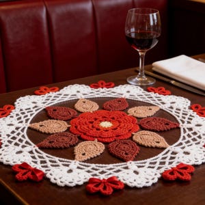May include: A round crochet doily featuring a central red flower surrounded by alternating light brown and dark brown leaf-shaped motifs. The outer edge is finished with a white lacy border accented by small red bows at regular intervals.