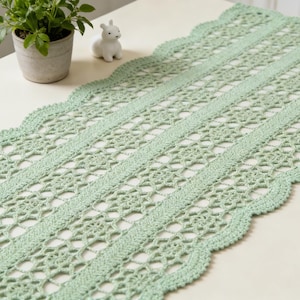以下が含まれることがあります： スカラップエッジの薄緑色の、かぎ針編みのテーブルランナー。ランナーは、長さに沿って走る無地のバンドと、繰り返しの透かし模様が特徴です。小さな鉢植えと白いセラミックのウサギが背景にあります。