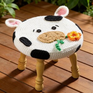 May include: A handcrafted, round stool with a cow design. The white crocheted top features black spots, a brown muzzle, and a flower. The stool has wooden legs and small pink ears. A whimsical and decorative piece.