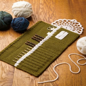 May include: An olive green crocheted roll-up case for knitting needles and crochet hooks, featuring a white lace trim, a white crocheted flap, and a small white pocket. The case holds several metal and wooden needles, accompanied by yarn balls.