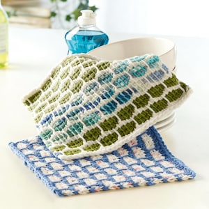 May include: Two handmade crochet dishcloths in the image. The top cloth features a green, white, and blue pattern, while the bottom cloth is blue and white. A stack of white plates and bottles of soap are in the background.