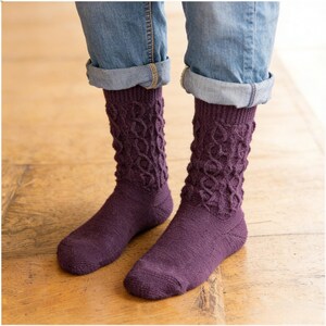 May include: A pair of dark purple knit socks with a cable knit pattern. The socks are worn with blue jeans with the cuffs rolled up, revealing the ankles. The socks are made of a soft, textured material.
