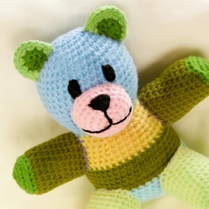 May include: A colourful, handmade crochet teddy bear. The bear has a blue head with green ears, a pink muzzle, and black stitched eyes and mouth. The body is striped with yellow and green. The arms and legs are green.