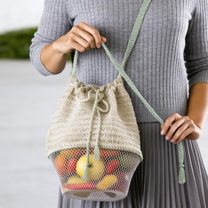 Pode incluir: Uma bolsa de balde de crochê bege com uma base de malha de metal, cheia de frutas coloridas. A bolsa tem um fecho de cordão e uma alça verde claro. A bolsa é segurada por uma pessoa que usa um suéter e uma saia cinza.