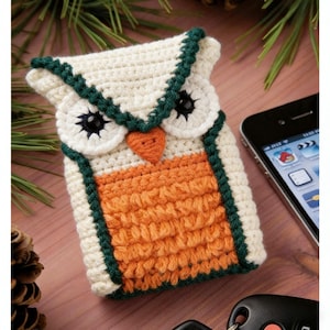 May include: A handmade crochet phone case shaped like an owl. The case features a cream-colored body with orange and green accents. The owl's eyes are black, and the beak is orange. The case is designed to fit a smartphone.